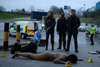 Drie detectives staan bij een plaats delict waar een dode man naakt op de grond ligt