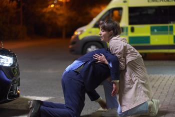 Twee mensen bij ambulance, één knielt, de ander biedt steun. Nachtelijke omgeving.