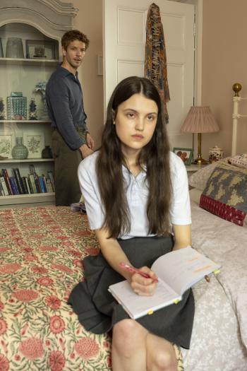 Jonge vrouw op bed met notitieboekje, man staat op de achtergrond in een slaapkamer.