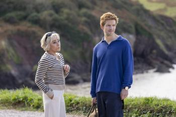 Twee personen staan buiten bij de kust, met groene heuvels op de achtergrond.