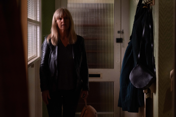 Vrouw met blond haar in donkere hal, draagt leren jas en tas.