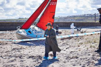 Vrouw in regenjas op strand, rode zeilboot op de achtergrond.