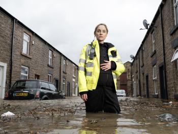 Vrouw in felgele veiligheidsjas staat in overstroomde straat tussen bakstenen huizen.