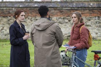 Twee vrouwen en een man praten buiten, naast een fiets en een stenen muur.