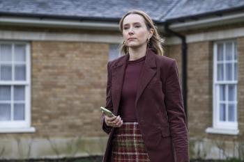 Vrouw in een bordeauxrode jas en geruite rok met telefoon voor een bakstenen huis.