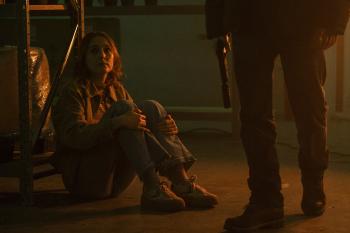 Vrouw zittend op de grond, kijkt angstig naar persoon met pistool.