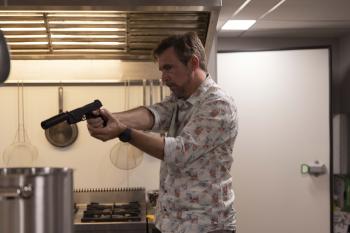 Man met geweer in een keuken, draagt een overhemd met patroon.