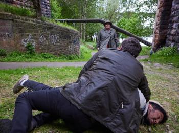 Twee mannen vechten op gras, derde man kijkt toe.