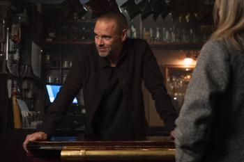 Man leunt op bar, kijkt naar persoon, sfeervolle baromgeving.