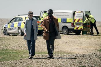 Twee mannen lopen over een strand, politieauto's en agenten op de achtergrond.
