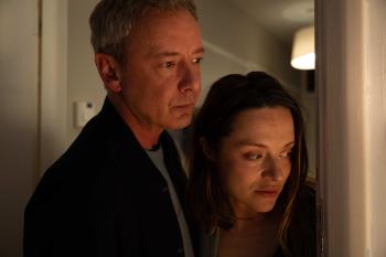 Man en vrouw kijken bezorgd door een deuropening in een schemerige kamer.