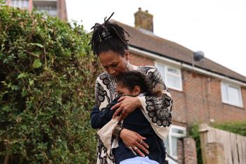 Vrouw knuffelt kind buiten bij huis, omgeven door groen.