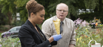Twee personen in een tuin, vrouw houdt een bewijszak vast, man kijkt verbaasd.