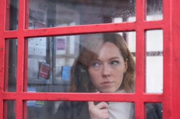 Vrouw aan de telefoon in een rode telefooncel, kijkt peinzend uit het raam.