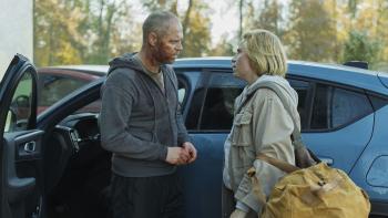 Een bebloede man en een vrouw voeren een intens gesprek naast een auto