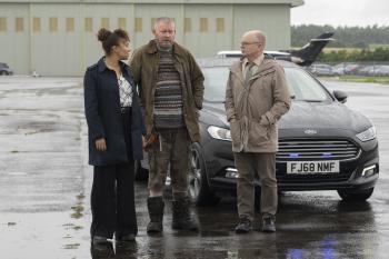 Drie mensen staan met een auto op een leeg terrein in de regen