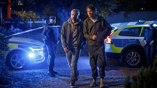 Twee mannen lopen op de plaats delict. Op de achtergrond zie je twee politieauto's en een politielint.