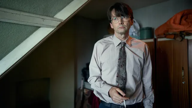 Man met overhemd aan en stropdas om en een sigaret in zijn hand staat in een kamer.