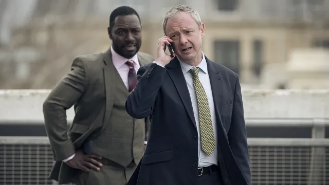 Detective Roy Grace en zijn partner staan allebei buiten in pak, Grace is aan de telefoon en beiden kijken bezorgd