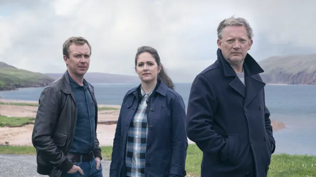 De hoofdpersonages van Shetland seizoen 6 staan met hun handen in hun zakken in de wind op de shetlandeilanden en kijken de camera in