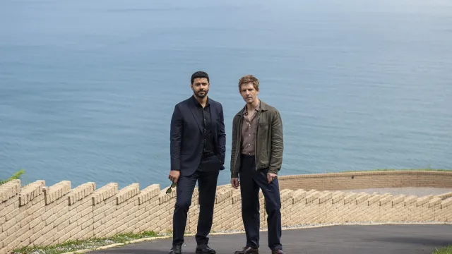 Twee mannen staan voor een zee met een weg en muur op de achtergrond.