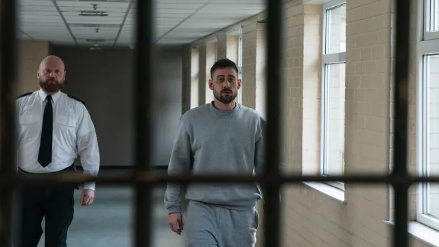 Mannelijke gevangene loopt naast bewaker in grijze gang.