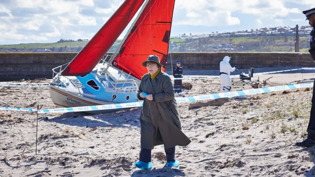 Vrouw in regenjas op strand, rode zeilboot op de achtergrond.