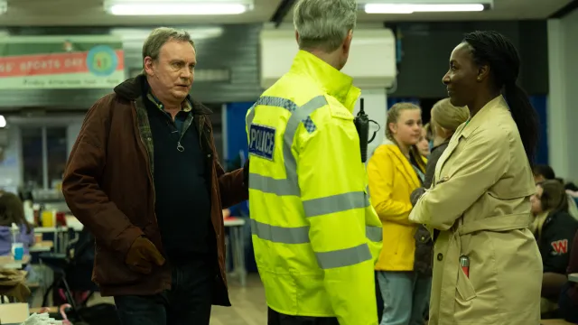 Mensen praten in een drukke zaal, een met politiejas en een met beige jas.