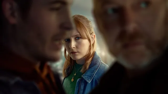 Een vrouw in een groene trui, kijkend tussen twee onscherpe mannen in de voorgrond.