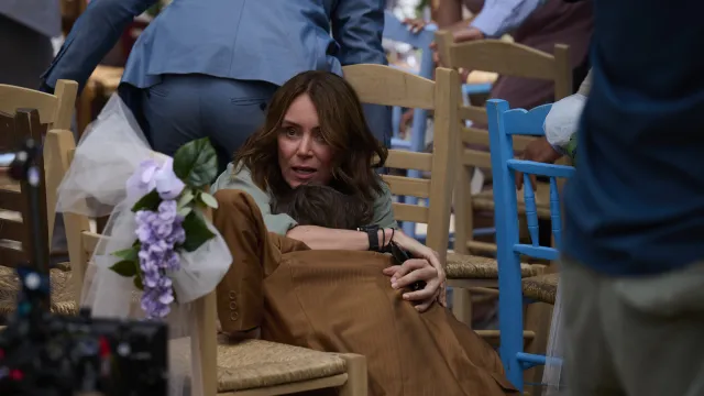 Een vrouw omarmt een kind zittend op een houten stoel, omringd door mensen en bloemen.