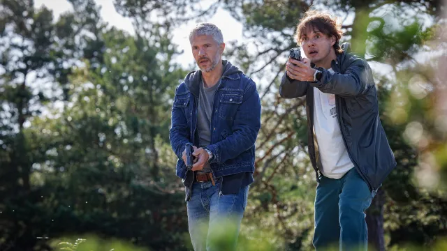 Twee mannen in een bos houden een pistool in de hand, alert kijkend.