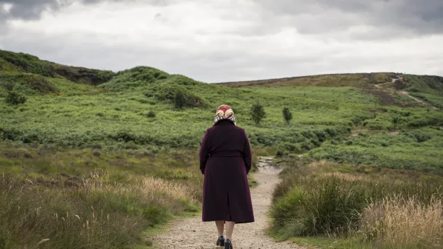 Een oude vrouw met rode jas loopt door een groen heuvellandschap