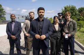 Zes mensen in formele kleding staan buiten, serieuze gezichtsuitdrukkingen.