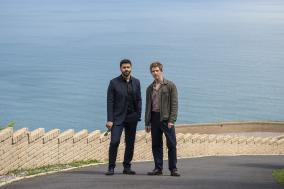 Twee mannen staan voor een zee met een weg en muur op de achtergrond.