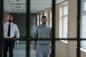 Mannelijke gevangene loopt naast bewaker in grijze gang.