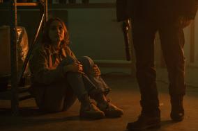 Vrouw zittend op de grond, kijkt angstig naar persoon met pistool.