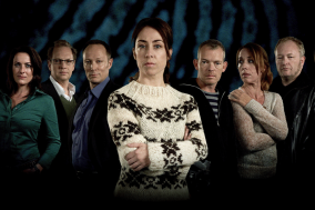 Groep mensen in donkere kleding, vrouw in lichte trui centraal, donkere achtergrond.