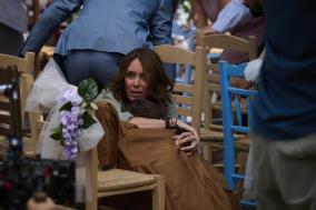 Een vrouw omarmt een kind zittend op een houten stoel, omringd door mensen en bloemen.