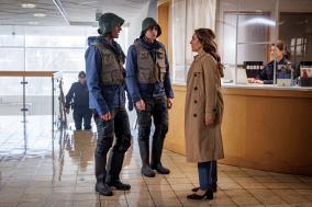 Twee agenten praten met vrouw in mantel bij balie in lobby.