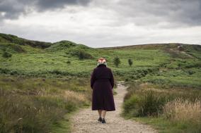 Een oude vrouw met rode jas loopt door een groen heuvellandschap
