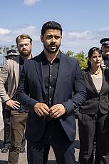 Zes mensen in formele kleding staan buiten, serieuze gezichtsuitdrukkingen.