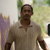 Man loopt buiten, draagt een beige gebreid shirt. Graffiti op de muur links.