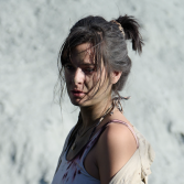 Vrouw met warrig haar kijkt somber, draagt een top met spaghettibandjes en sjaal, lichtgrijze achtergrond.