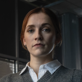 Vrouw met bruin haar in een kantooromgeving, serieus kijkend.