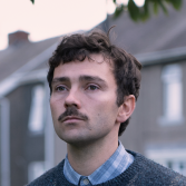 Man met snor kijkt serieus, grijs huis op achtergrond.