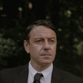 Mannelijke acteur in donker pak, buiten in groene omgeving.